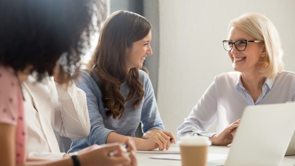 Friendly smiling old and young businesswomen talking laughing at corporate group business meeting, happy female employees colleagues coach manager and intern having fun pleasant conversation at work lobdock advantage